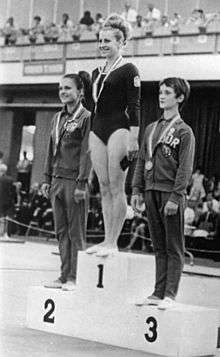 Three young women standing on a sportspodium with medals around their necks. The winner, standing in the middle on the top, is wearing a gymnastics dress with a little lion on her left arm. The silver and bronze medalists, standing on the sides, are wearing casual sports clothes with a sign "DDR". The winner and the silver medalist are smiling happily, the bronze medalist looks serious.