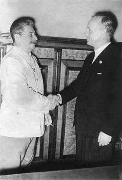 Two smiling men shake hands. Man on left wears a white suit and holds a cigarette. Man on right wears a black suit.