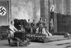 A black-and-white photo of six soldiers standing around a flag-covered coffin. All six soldiers wear steel helmets and numerous military decorations. To the left of the coffin is a lectern with a man in a military uniform speaking into two microphones on the lectern. The wall behind the coffin and lectern is draped with a swastika flag. To the right of the lectern is a pillar with an iron cross on the front and an eagle sitting on top.