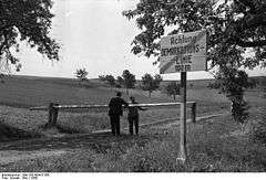 Two people standing either side of a lowered border pole on a dirt road with a sign in the foreground