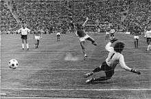 A black-and-white photograph of footballers in action, taken from behind the goal as a penalty kick is taken. The ball has been kicked by the taker to the left of the goalkeeper, who has dived forlornly to his right.