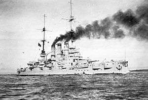 A large battleship cruises slowly near the coast. Wind blows the smoke emitted from the three funnels over the ship