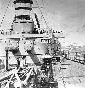 Two long gun barrels jut out of a wide, round gun turret aboard a warship; the deck is covered in heavy chains, winches, and cranes
