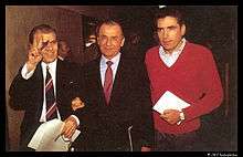Three men are walking side-by-side holding papers. The first two are wearing a suit and the third is wearing a red sweater. The first man is smiling and flashing a V sign.