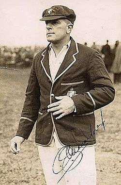 Stocky man in white cricket shirt and trousers with a blazer with the Australian coat of arms over the top of the shirt. His left hand is on his stomach and his right fingers are pressing something together, possibly a cigarette. The photo is signed by Macartney and there is a crowd of people in the background, and grass can be seen.