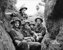 Five young Caucasian men in uniform wearing helmets sit below ground in a trench, facing the camera. The trench is central to the photograph and runs away from it, with the edges of the earthworks on each side. In the background, the skyline can be seen to the rear of the men, framed by the edges of the trench which are covered in roots, grass and other vegetation.