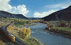 View of a light blue river flowing through a forested valley, with a train running alongside at left and snowcapped peaks in the background