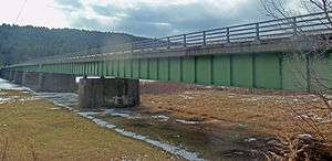 A concrete bridge with green undergirding and no overhead spans a river at left and land with some snow on it below at right