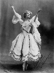 A woman in a white ballet skirt stands on the tips of her toes, arms extended upwards