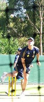 A man wearing dark blue cricket training kit with gold piping bowls a cricket ball. A net can be seen predominately in the fore-ground.