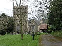 Square gray tower of stone church building, partially obscured by trees. Red roofed lych gate to right. Grass and gravestones in the foreground