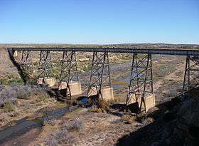 A small stream in arid country flows under a railroad bridge high above the water.
