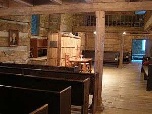 Photograph of the interior of an old log church with a rough hewn timber supporting column near the center of the image. The column supports a timber beam. Other beams are visible supporting a balcony that surrounds the room on three sides. The photograph is facing towards a communion table at the front of the church, and is taken from the left side of the room beneath the balcony. Plane wooden pews are visible to the left and on the other side of the room. The floor is wooden. A portrait of Thomas Campbell is visible to the left, on the front wall of the room.