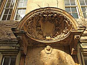 A large decorated shell-like stone canopy, with a shield in the centre and carved foliage around; a cherub's head underneath the hood