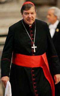 George Cardinal Pell wearing the ordinary dress of a cardinal: black cassock with scarlet (red) piping and buttons, scarlet fascia (sash), pectoral cross on a chain, and a scarlet zucchetto.