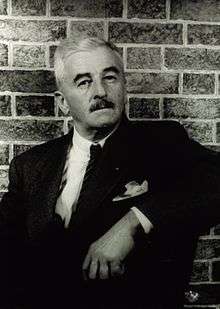 An old man, dressed smartly in a suit and tie, rests his left arm on the arm of a chair. Close behind him is a simple brick wall.