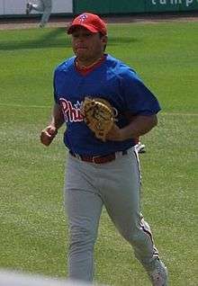 A man in a blue baseball jersey, white baseball pants with red pinstripes, and a red baseball cap jogs across the baseball field wearing a tan catcher's mitt on his left hand.
