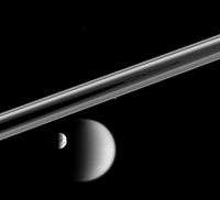 Circular complex rings of Saturn are seen at the low angle. The rings look like two grayish bands running parallel to each other from the left to right and connecting at the far right. Half illuminated Titan and Dione are visible slightly below the rings in the foreground. Two bright dots: one at the lower edge of rings and another above the rings can be seen. They are Prometheus and Telepso.