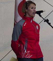 A woman with short blonde hair speaks into a microphone to an unseen audience. She is wearing a red and grey coat.