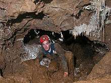 A man caving in muddy passage with helictite formations on the walls and ceiling