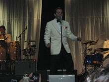 A man wearing a white tuxedo performing at a concert.