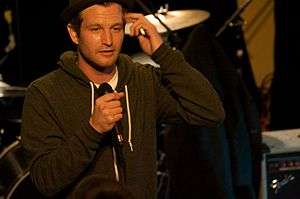 A man in a gray hoodie and hat scratches his head with his left hand and grasps a microphone with his right