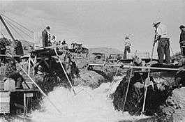Four men dressed in long-sleeved shirts, long pants, and hats are perched on platforms on both sides of a rushing stream. Three of the men are standing, and one is seated. Each man holds one end of a long pole with a net, dipped in the water, attached to the other end. Several people without poles are watching or waiting nearby.