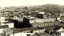 Photograph from an elevated vantage point looking over the rooftops of multistory buildings in a crowded downtown district toward a large bay with many steam and sailing ships riding at anchor and a mountain in the distance on the other side of the inlet