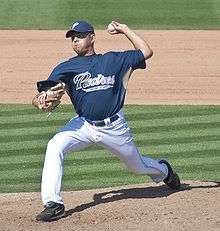 A man in white pants, a blue baseball jersey with "PADRES" in gold on the chest, a blue baseball cap, and a tan baseball glove on his right hand prepares to pitch a baseball with his left hand.