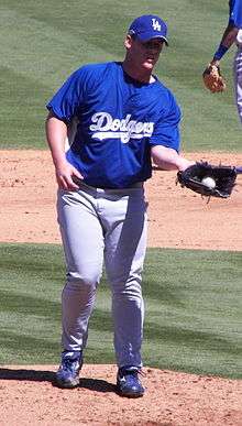 A man in a blue baseball jersey, cap, and gray pants catches a baseball in a glove on his left hand.