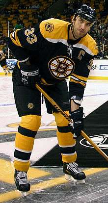 Zdeno Chara posting for a picture on the ice with his hockey stick as a Boston Bruins team member.