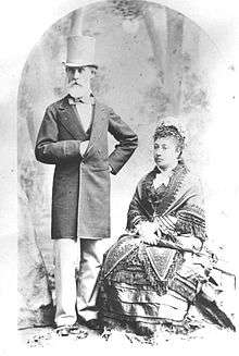 An old man in a top hat with a white beard stands next to a sitting woman in a decorative dress