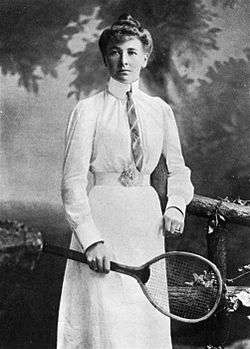 A black and white picture, a woman is in all-white attire with a tie on, and is looking right at the camera in the photograph with a racket in her right hand