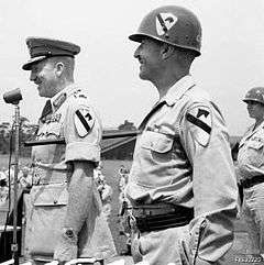 Two soldiers stand at attention. The one on the right wears an American uniform and helmet bearing the patch of the 1st Cavalry Division. The one on the left wears an Australian uniform with peaked cap and generals' gorget patches and carries a swagger stick, but incongruously also wears the patch of the 1st Cavalry Division on his sleeve.