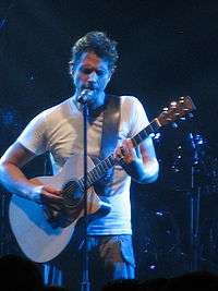 A man wearing a white T-shirt, playing a guitar with his eyes closed while standing behind a microphone stand.