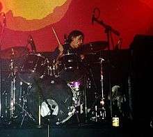 A man performing on a purple drum kit against a red-and-yellow backdrop. A drum stick is visible in his right hand; several microphones and a cooling fan are also visible.