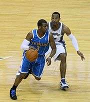 A black basketball player, wearing a blue jersey with a word "New Orleans" and the number "3" written in the front, dribbles the ball in front of another player
