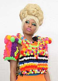 An Afro-American woman in a blonde wig and bright teal eyeshadow wears a shirt constructed of variously colored cotton balls