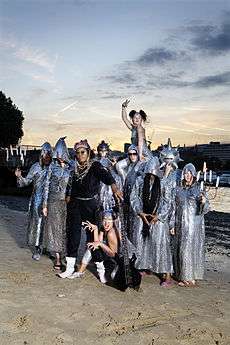 Chrome Hoof on a dawn/dusk Summer of 2007 river beach, dressed to promote 'Pre-Emptive False Rapture'.  Flanking members hold candelabra and most are wearing silvery cowls, with Shingai Shoniwa counterpointed in matt black.
