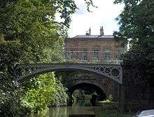 A iron bridge spanning water. In the background is a yellow stone building. On the left trees reach out over the water.