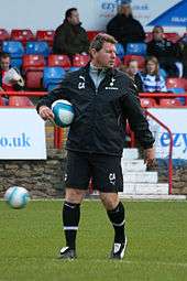 A football coach wearing all black, stands on a football pitch holding a football.