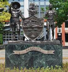 A bronze statue is seen that depicts most of the details of the coat of arms shown in the infobox image. The men and the sloop are three-dimensional representations of the respective components in the shield. The shield itself is relief work.