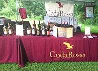 A table with a red tablecloth marked "Coda Rossa" and bottles of wine, which is under a tent with woods in the background.