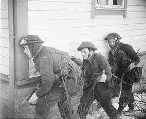 three British soldiers take cover at the corner of a house