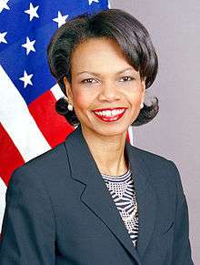 Condoleezza Rice smiling with thickly applied red lipstick wearing a dark blue jacket over a patterned blouse. The United States flag is in the background.