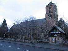 A sandstone church seem from the northwest; the tower to the right has a small pyramidal spire. In the foreground is a wall and a lych gate with a banner saying "Christmas Joy to All"