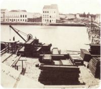 An old photograph showing piles of construction materials and equipment along the bank of a river with large white buildings lining the opposite bank