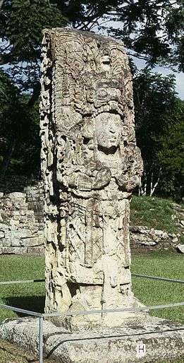 Intricately carved free standing stone shaft sculpted in the three-dimensional form of a richly dressed human figure, standing in an open grassy area.