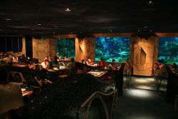 A photograph of the interior of a dimly lit restaurant where people are eating next to three windows into an aquarium housing a variety of marine organisms