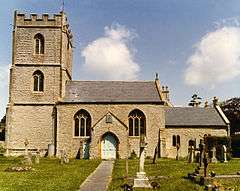 Stone building with square tower at left hand end. In the foreground either side of a path are gravestones in a grassy area.